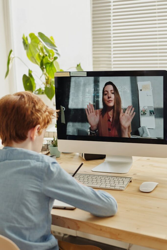 A child participates in online learning through a video call with a teacher, using a desktop setup indoors.